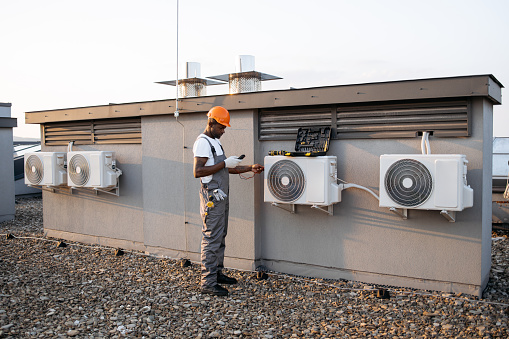 Full length view of busy foreman standing near white air conditioner with toolbox on it. Skilled african american male using digital multimeter for testing power in factory equipment.