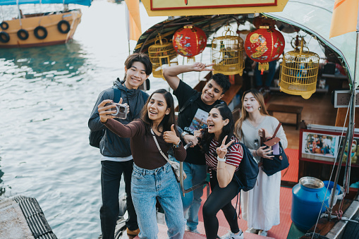 A wide shot of a young female tourist taking a selfie photo on her phone with a group of other tourists of mixed nationalities. They are smiling and posing for a photo. The tourists are standing on a small boat in the Aberdeen's fishing port