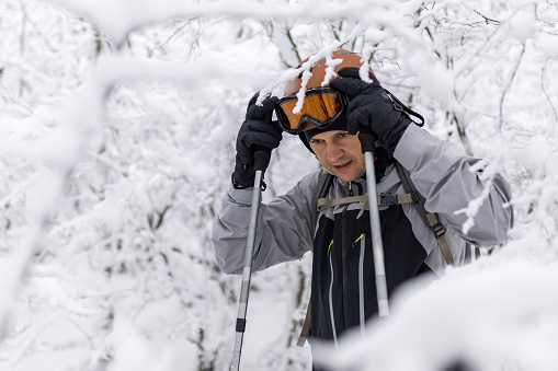 Male hiker with trekking poles adjusting ski googles while walking among trees on snow mountain