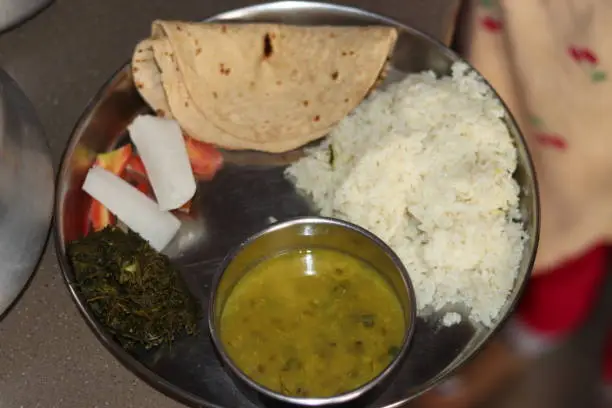Photo of A steel plate thali with Normal Indian Staple food including radish, carrot, rice, chapati, daal, gajak, selectively focus