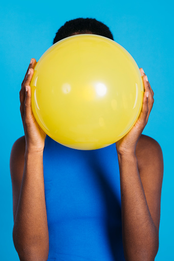 Woman blowing a yellow balloon in front of blue background in a studio. Woman with short hair inflating a yellow balloon.