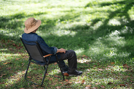 Back of the relaxed senior Asian man sits comfortably in an outdoor chair, surrounded by the soothing beauty of nature