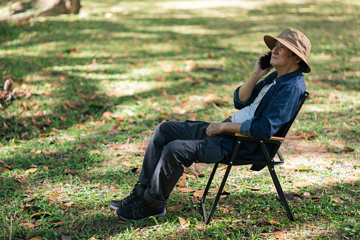 Senior Asian man seated on an outdoor chair, using a smartphone, with a serene nature background