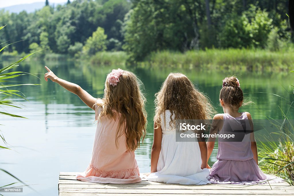 Three girl friends together on river jetty. Three young kids sitting together on jetty at lakeside. Sitting Stock Photo