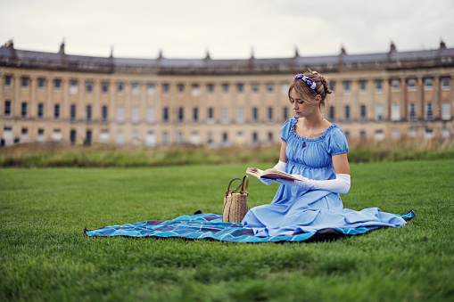 Young woman wearing a regency era dress enjoying reading a book in front of Royal Crescent building in Bath, Somerset, United Kingdom.\nShot with Canon R5