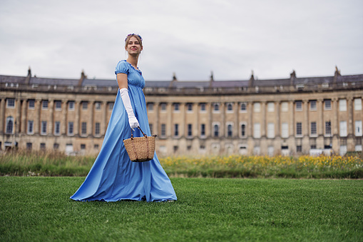 Young woman wearing a regency era dress enjoying a walk in front of Royal Crescent building in Bath, Somerset, United Kingdom. The girl is holding a small basket.\nShot with Canon R5