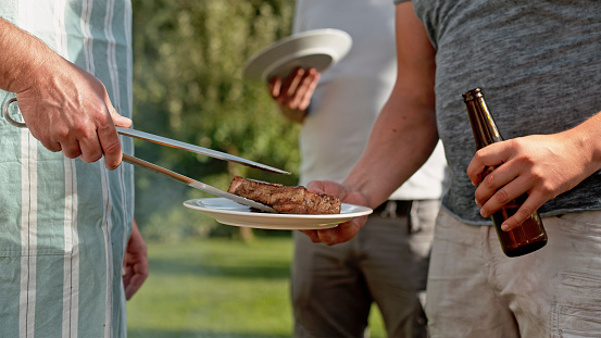 Man taking food from charcoal grill at garden party and placing it onto plates of guests.