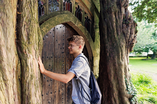 Teenage boy is sightseeing Stow-on-the-Wold, a beautiful village in Cotswolds, Gloucestershire, United Kingdom. He is admiring beautiful church door and trees standing on it's both sides.
Shot with Canon R5