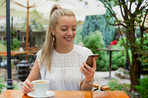 Young woman enjoying a cup of coffee and using her smart phone sitting outdoors