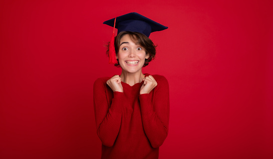 It's cool to have a degree. Portrait of beautiful funny young graduate girl with fists up isolated in studio on red background. University, study, education, college, knowledge, graduation