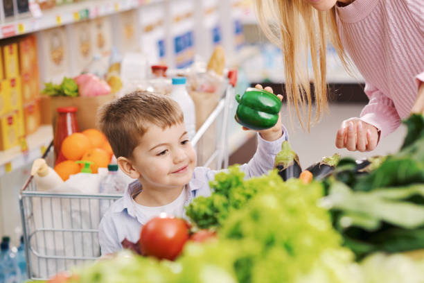 Smiling caring mother and cute kid doing grocery shopping together, they are picking vegetables in the produce section at the supermarket