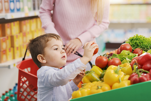 Cute smiling boy with his mother at the supermarket, he is picking fresh vegetables: grocery shopping and healthy eating concept