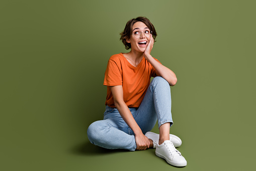 Full size photo of impressed woman dressed orange t-shirt jeans in glasses sit look at sale empty space isolated on khaki color background.