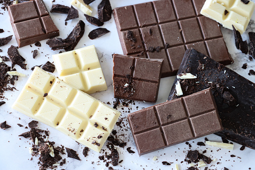 Stock photo showing close-up, elevated view of bars of milk, white and dark chocolate. The milk chocolate has been broken down into chunks, the white chocolate in squares, whilst the dark chocolate has been grated into shavings.