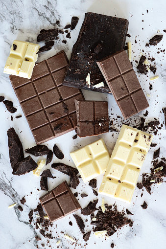 Stock photo showing close-up, elevated view of bars of milk, white and dark chocolate. The milk chocolate has been broken down into chunks, the white chocolate in squares, whilst the dark chocolate has been grated into shavings.