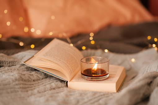 Burning scented candle on open paper book over lights in bed close up. Aromatherapy.