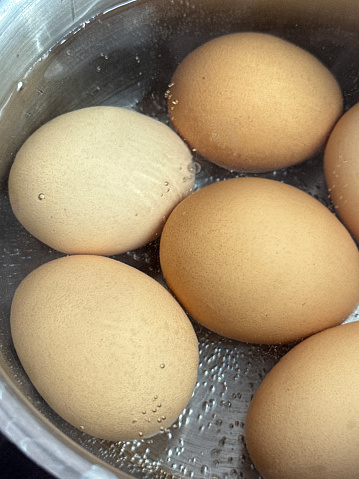 Stock photo showing close-up, elevated view pf six chicken eggs being boiled on a hob, in a stainless steel saucepan of boiling water. These are organic, free range chicken eggs, which vary greatly in size, shape and colour.