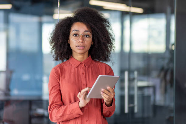 istock Serious confident woman in red shirt inside office with tablet computer in hands, looking at camera in concentration and thinking, businesswoman at workplace inside office, programmer developer 1872234723