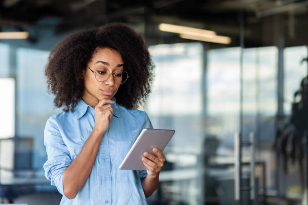istock Serious thinking woman standing near window inside office, businesswoman using tablet computer, reading data with focus and concentration, female programmer in glasses and shirt testing programs 1872234879