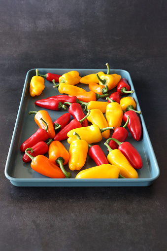 Stock photo showing close-up, elevated view of cooking tray covered in mini red, orange and yellow bell peppers (Capsicum annuum) on black  background.