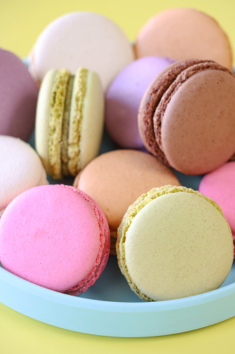 Stock photo showing close-up view of a plate of multi coloured macarons on a yellow background. Pink strawberry, white vanilla, purple blueberry, green pistachio, brown chocolate and orange flavoured macarons.