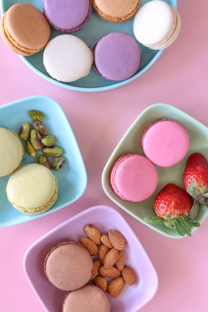 Image of plate of multi coloured macarons filled with flavoured butter cream, strawberry, vanilla, blueberry, orange, pistachio, chocolate flavoured meringues, strawberries, almonds and pistachio nuts, pink background, elevated view