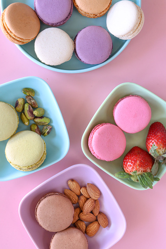 Stock photo showing close-up view of a serving plate of multi coloured macarons on a pink background. Pink strawberry meringues are served on a plate with strawberries, green pistachio with pistachio nuts and brown chocolate with almonds.