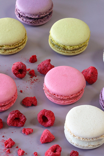 Stock photo showing close-up view of a batch of multi coloured macarons surrounded by dehydrated raspberries on a purple background. Pink strawberry, white vanilla, purple blueberry and green pistachio flavoured meringues.