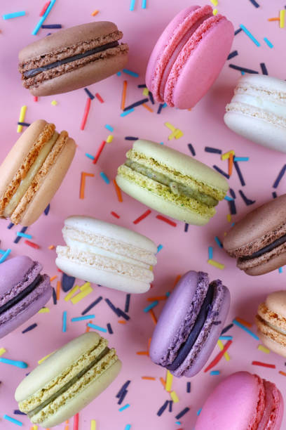 Full frame image of group of multi coloured macarons filled with flavoured butter cream standing on edge, strawberry, vanilla, blueberry, orange, pistachio, chocolate flavoured meringues, spilled sugar sprinkles, purple background, elevated view