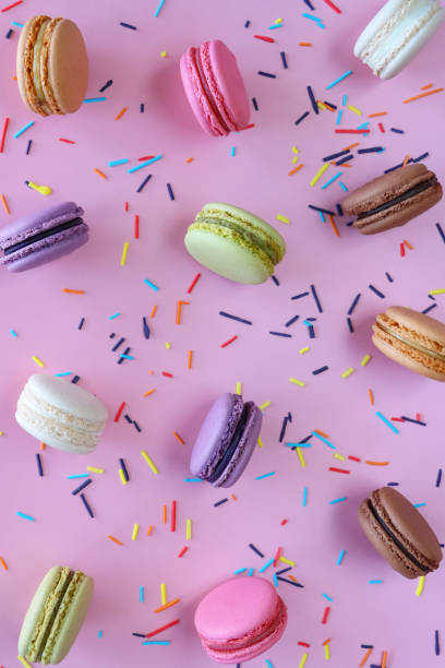 istock Full frame image of group of multi coloured macarons filled with flavoured butter cream standing on edge, strawberry, vanilla, blueberry, orange, pistachio, chocolate flavoured meringues, spilled sugar sprinkles, purple background, elevated view 1872259729