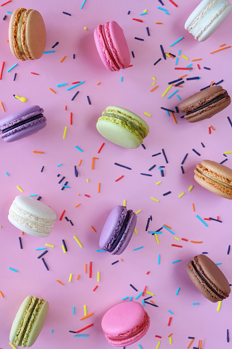 Stock photo showing close-up, elevated view of a batch of multi coloured macarons surrounded by spilled rainbow coloured sugar strand sprinkle on a purple background. Pink strawberry, white vanilla, purple blueberry, green pistachio, brown chocolate and orange flavoured meringues.