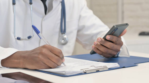 istock Close up of African Doctor Using Smartphone while Doing Paperwork in Hospital 1872294747