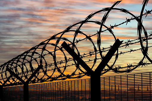 Security fencing with coiled barbed wire silhouetted against a colorful sunset, symbolizing border security and protection.