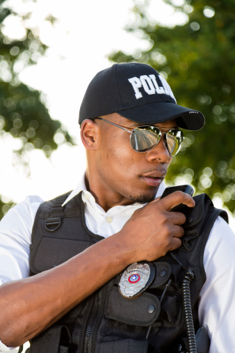 Policeman of African descent calls in a crime on his radio.  He wears bulletproof vest and mirrored sunglasses.  