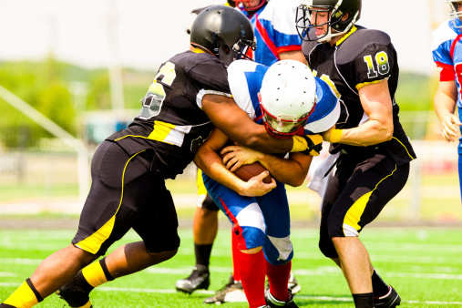 A football player is running with the ball in his arms.  He is holding the ball tight, while two players from the other team are trying to take it.  The man with the ball is dressed in a blue and white uniform.  The other team's uniform is black and yellow.