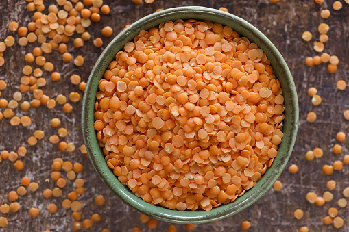 Stock photo showing close-up, elevated view of split red lentils that are piled high in a green, china bowl against a distressed brown background. Lentils are considered to be a very healthy food, containing a rich source of wealth of carbohydrates and proteins, as well as B vitamins, calcium, iron and phosphorus.