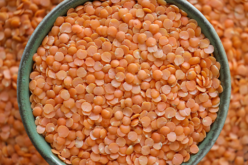 Stock photo showing close-up, elevated view of split red lentils that are piled high in a green, china bowl against a lentil layer background. Lentils are considered to be a very healthy food, containing a rich source of wealth of carbohydrates and proteins, as well as B vitamins, calcium, iron and phosphorus.