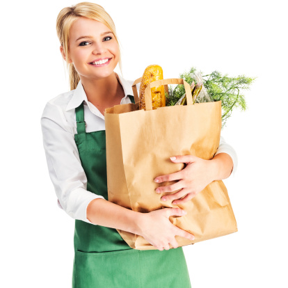 Photo of a young woman in a green apron carrying a paper bag full of groceries; isolated on white.