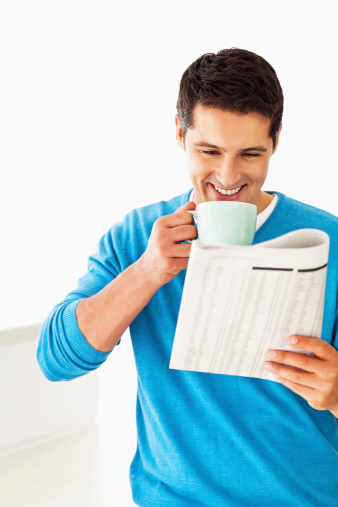 Happy young man in sweatshirt having coffee while reading newspaper. Vertical shot.