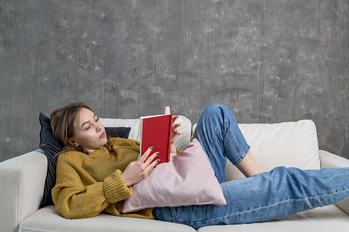 young woman is reading a red book on the sofa. a girl in a yellow sweater and blue jeans