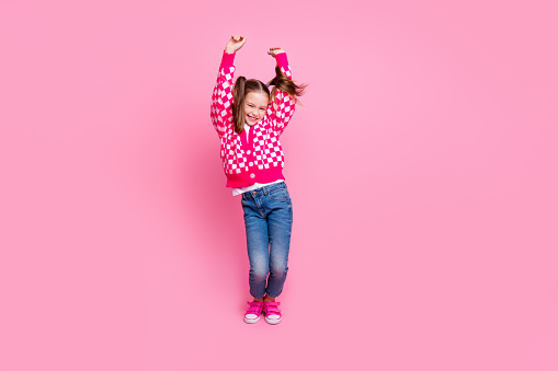 Portrait of lovely funky girl with ponytails wear print sweater denim pants raising hands up dancing isolated on pink color background.