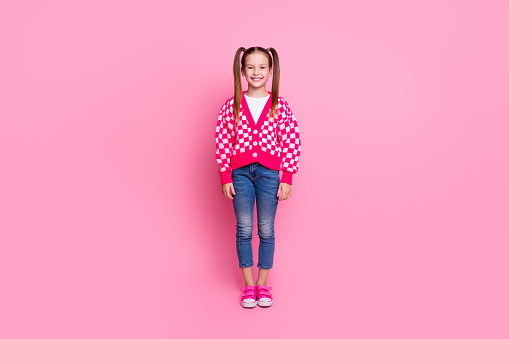 Full length photo of clever positive nice girl with ponytails dressed cardigan denim pants standing isolated on pink color background.