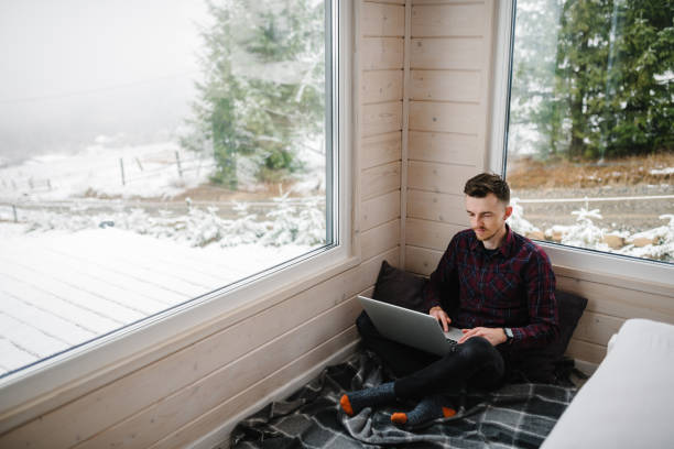 istock Remote work from cozy place. Man working on laptop in front of panoramic window with great view of mountains. Workplace in house in snowy morning. Wide interior view. Concept freelance lifestyle. 1872540434