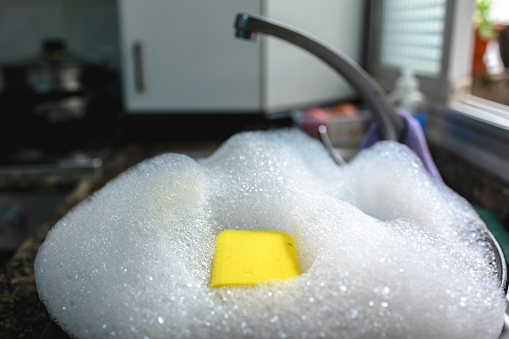 This close-up image captures a yellow sponge adrift in a sea of soap suds filling the kitchen sink, with a glint of sunlight reflecting off the watery foam. The sponge sits at the center, surrounded by glistening bubbles, with the blurred backdrop of a well-lit kitchen and window suggesting a domestic setting. The scene conveys a sense of pause in the midst of kitchen cleaning, highlighting the small and often overlooked elements of daily life.