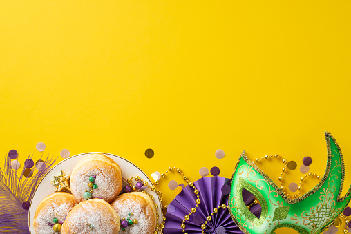 Extravagant Revelry Composition: Overhead shot showcasing table adorned with carnival mask, feather, fan, confetti, beads garland, donuts. Lively yellow background provides ideal space for text or ads