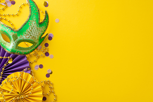 Festive Finery Arrangement: Top view capturing extravagant New Orleans mask, colorful bead strand, confetti, and decorative paper fans on a lively yellow background with space for messaging