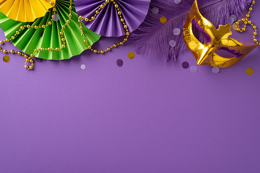 Festive Finery Arrangement: Top view capturing an extravagant New Orleans mask, colorful bead strand, feathers, confetti, and decorative paper fans on lively violet background with space for messaging