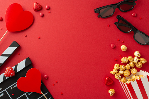 Top view romantic tableau featuring slate, 3D spectacles, popcorn box, delightful chocolates, sprinkles, heart-adorned sticks on vibrant red setting. Perfect for celebrating love and movie magic