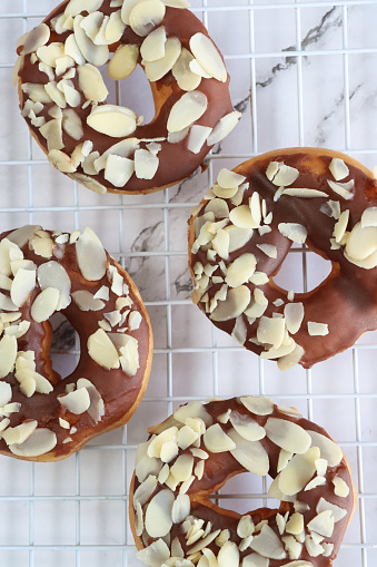 Stock photo showing close-up, elevated view of batch of chocolate glazed ring doughnuts topped with flaked almonds, on white cooling rack over marble effect background.