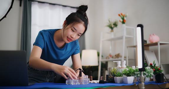 Young asian female photographer with side hustle Use laptop computer and camera, creativity her home studio as she arranges products and beauty props for photo shoot.